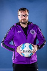 Abteilungsleiter Fußball_klein Mann in lila Sportjacke hält einen Fußball in den Händen.