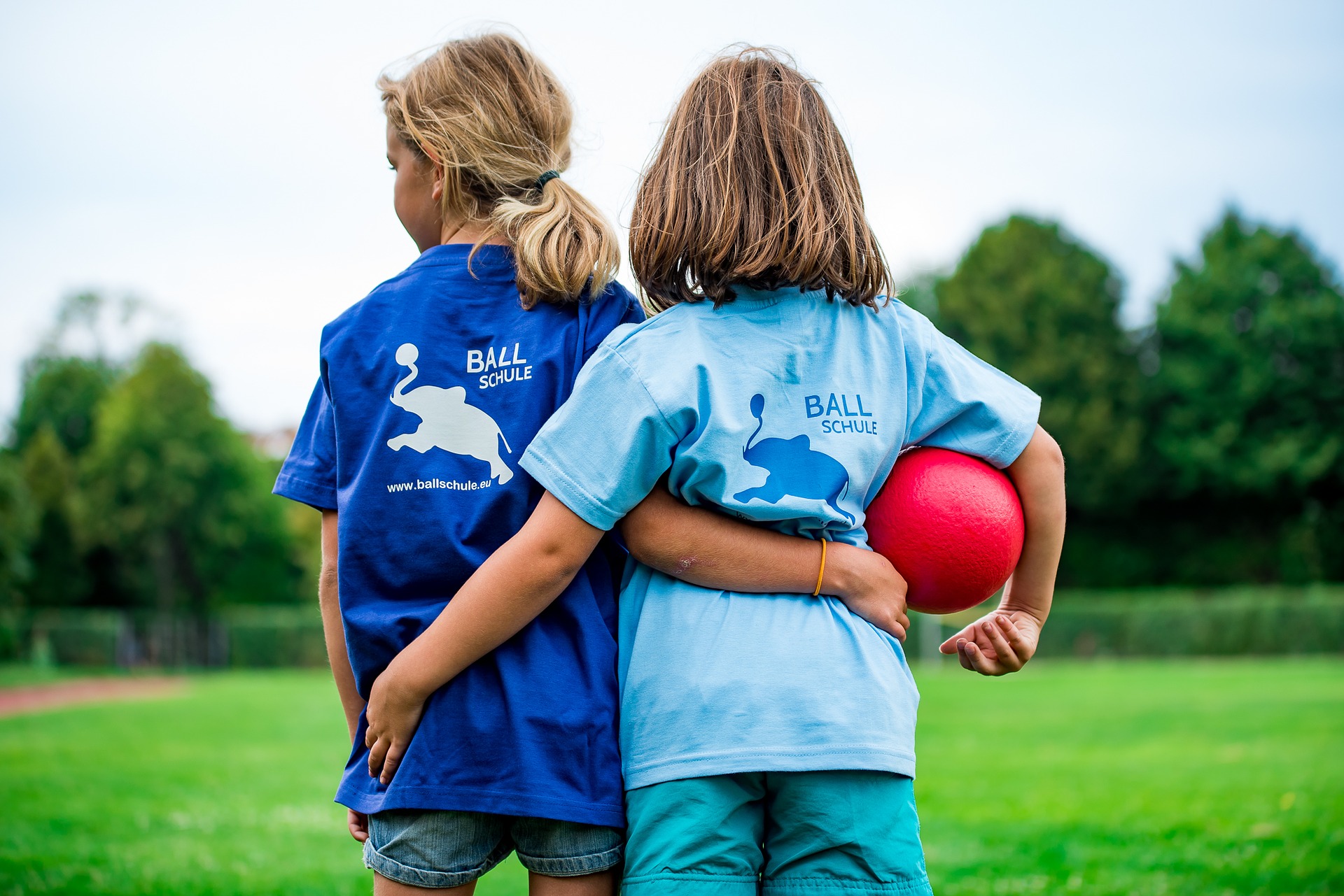 Handball Kinder Zwei Kinder in T-Shirts mit "BALLSCHULE"-Aufdruck, eines hält einen roten Ball.