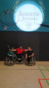 Drei Rollstuhlfahrer posieren in einer Sporthalle vor einem Badminton-Logo.