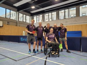 WhatsApp Image 2023-09-12 at 21.20.33(1) Sechs Personen in Sportkleidung stehen in einer Tischtennishalle, eine Person im Rollstuhl hält einen Tischtennisball.