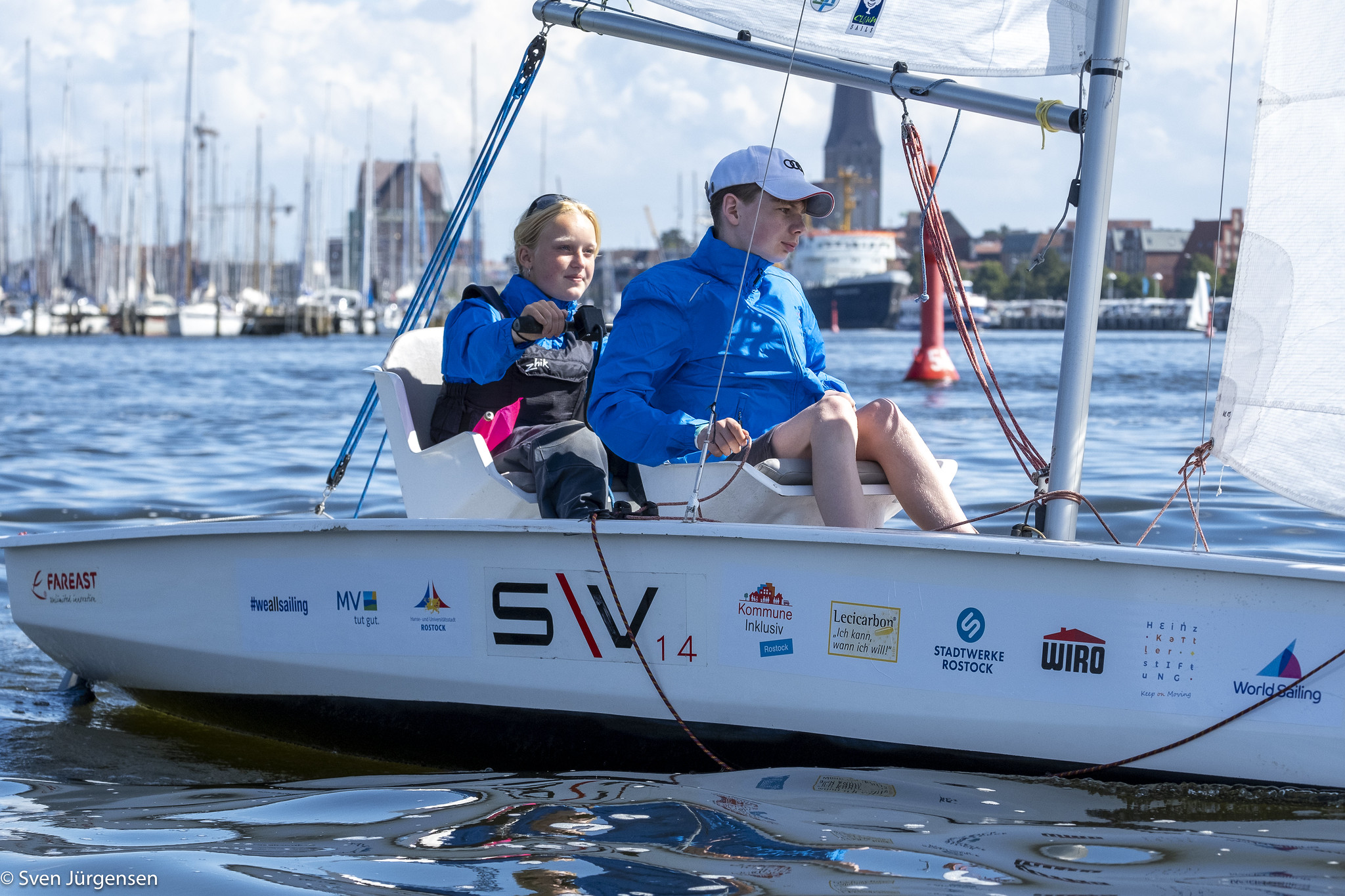 Zwei Kinder in Segelboot auf dem Wasser, umgeben von Hafenanlagen.