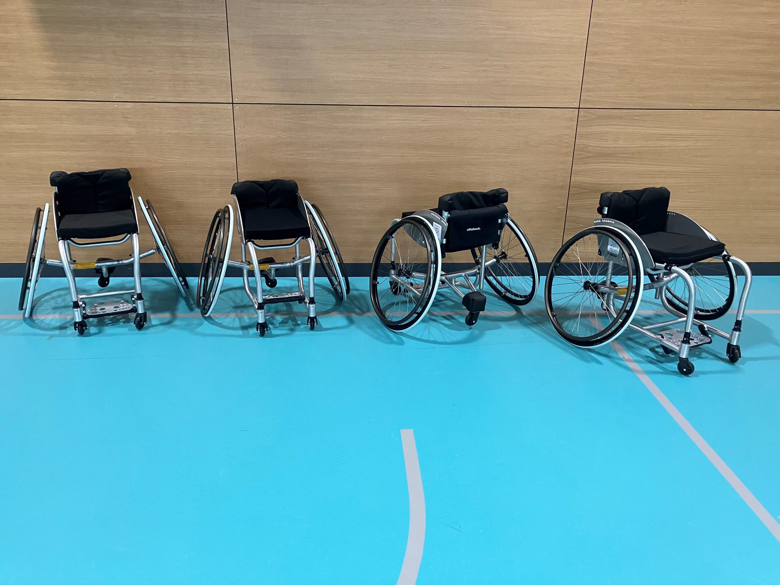 Vier Rollstühle auf einem blauen Sportboden.