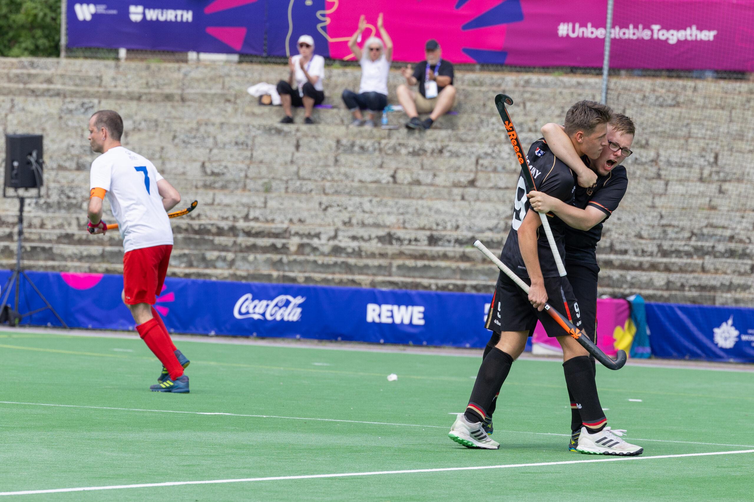 Zwei Hockeyspieler umarmen sich freudig auf dem Spielfeld, Zuschauer applaudieren im Hintergrund.