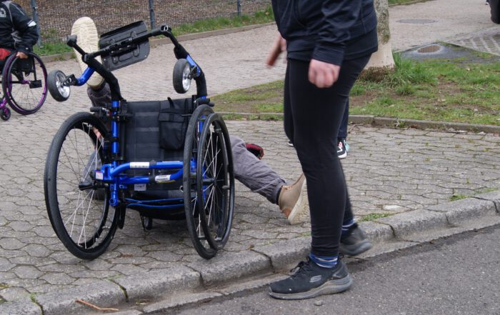 Rollifahrer liegt rücklings auf dem Biden, neben ihm ein Trainer, sie übern das aufstehen, nachdem man umgefallen ist