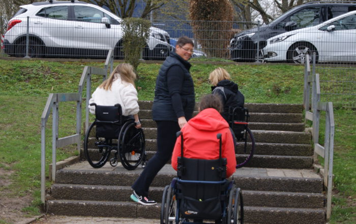 3 Rollstuhlfahrer auf und an einer Treppe mit mehreren Stufen. Trainer steht unterstützend zur Seite