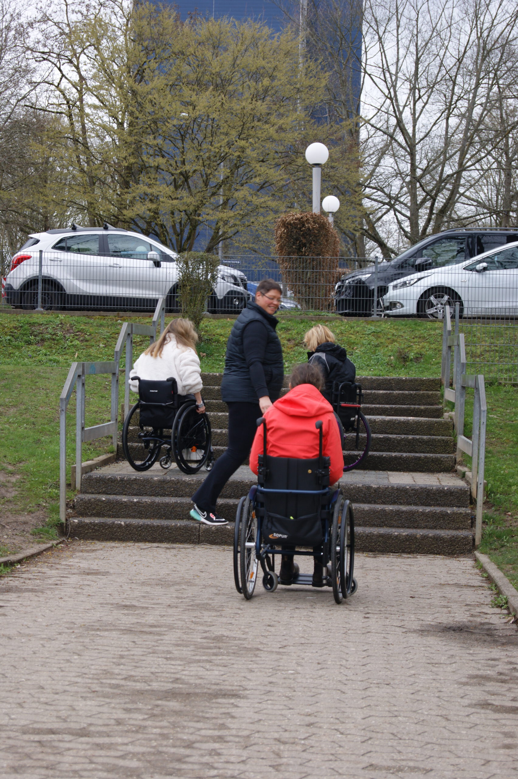 3 Rollstuhlfahrer auf und an einer Treppe mit mehreren Stufen. Trainer steht unterstützend zur Seite