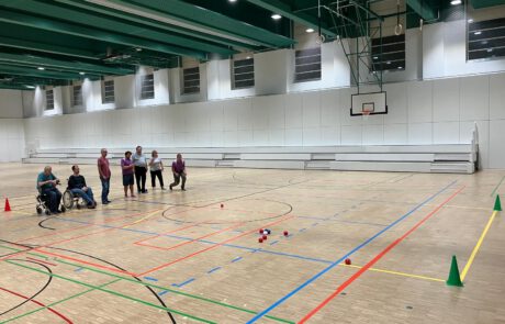 Para-Boccia-Spielszene in einer Sporthalle: Athletinnen und Athleten beobachten den Wurf, im Vordergrund Boccia-Bälle auf dem Spielfeld