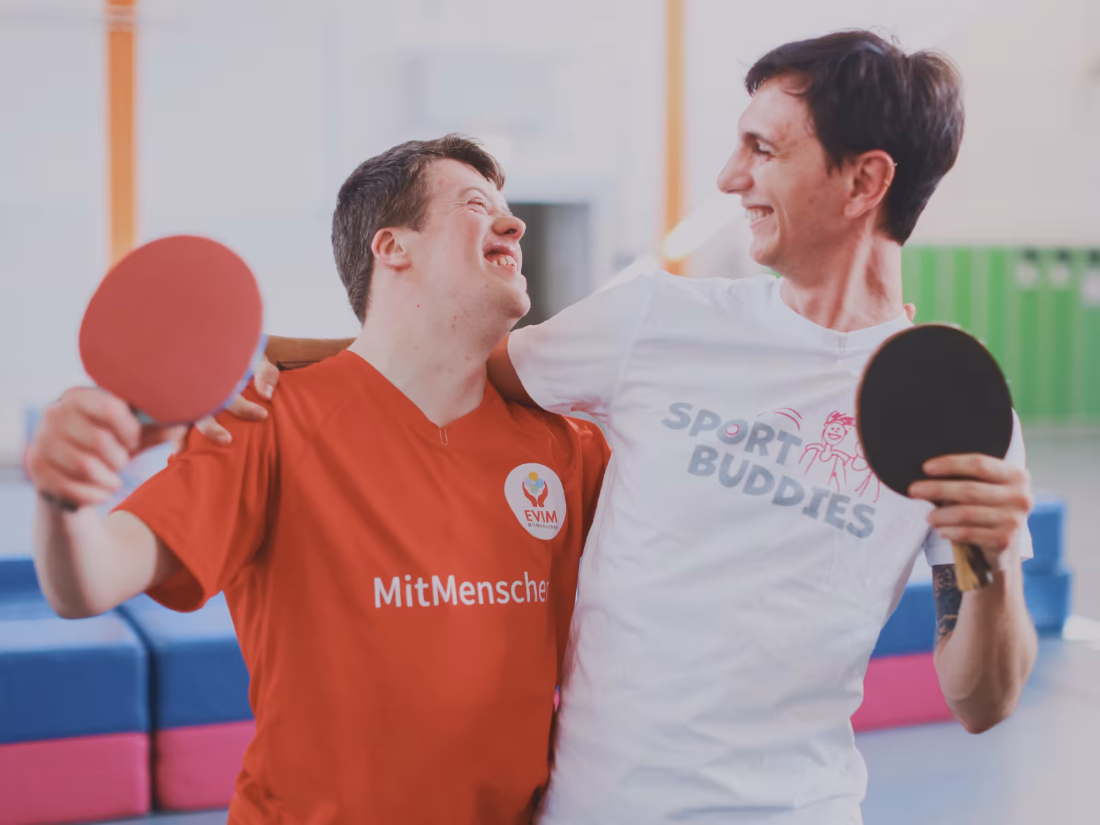Zwei Sportbuddies stehen mit Tischtennisschlägern im Arm und lachen sich an.