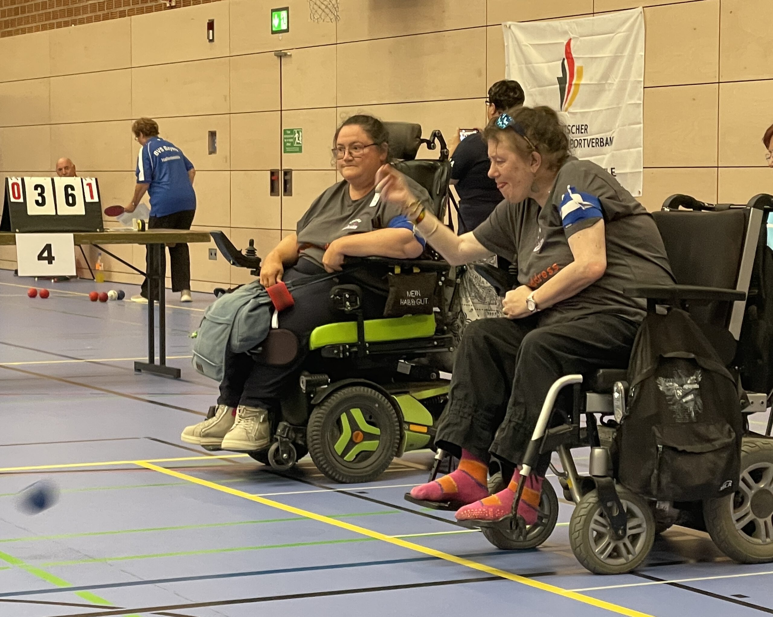 Zwei Spielerinnen der Boccia Rhinos in Rollstühlen während eines Spielzugs, im Hintergrund die Anzeigetafel mit dem Spielstand.